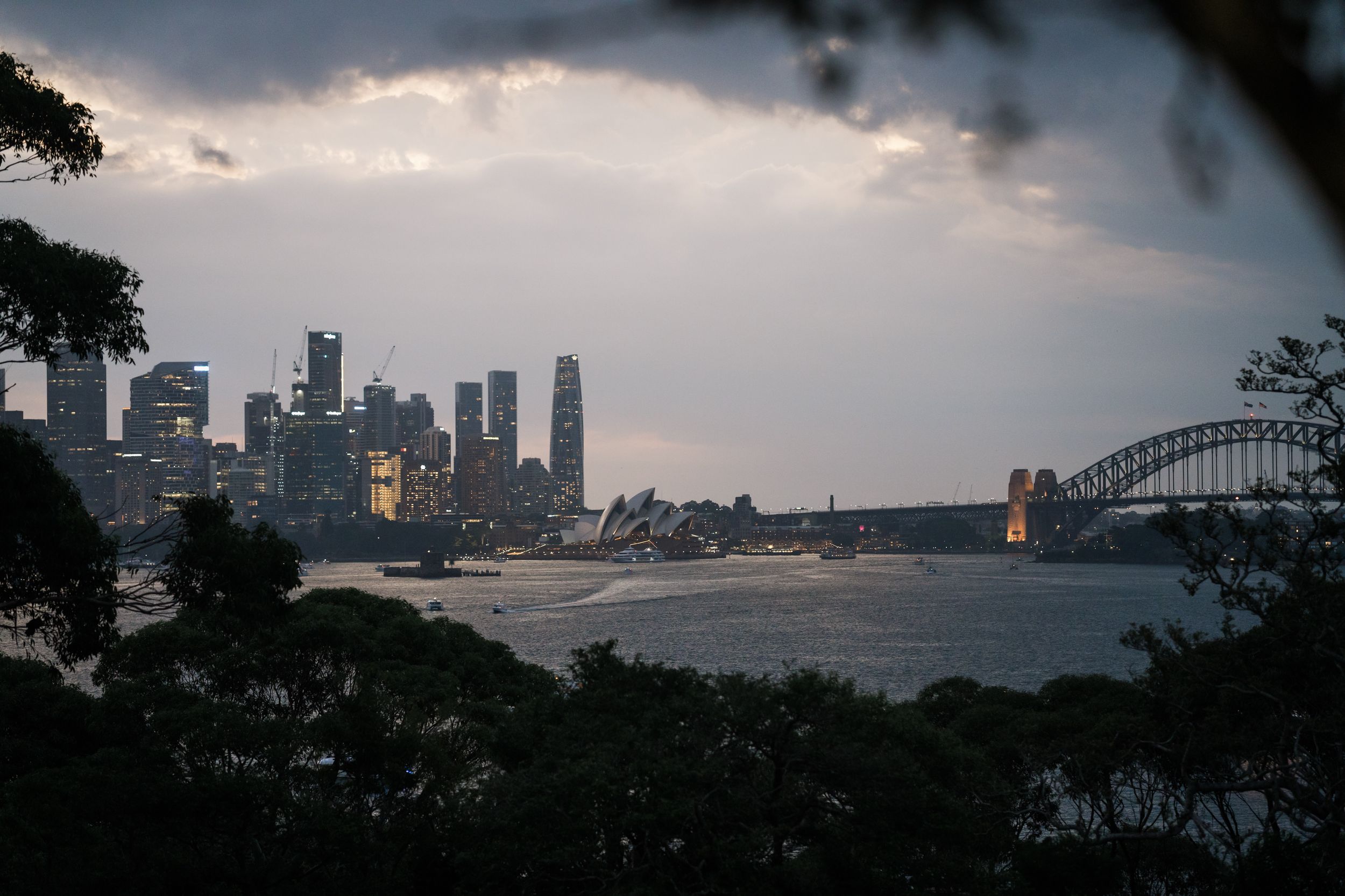 Sydney Harbour skyline at dusk with Opera House, Harbour Bridge, and stormy clouds over calm waters.