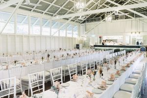 Elegant wedding reception hall with long white tables, chiavari chairs, and floral centerpieces under a bright skylit roof.