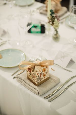 Elegant wedding table setting with a floral gift box tied with a gold ribbon on a linen napkin beside silverware.