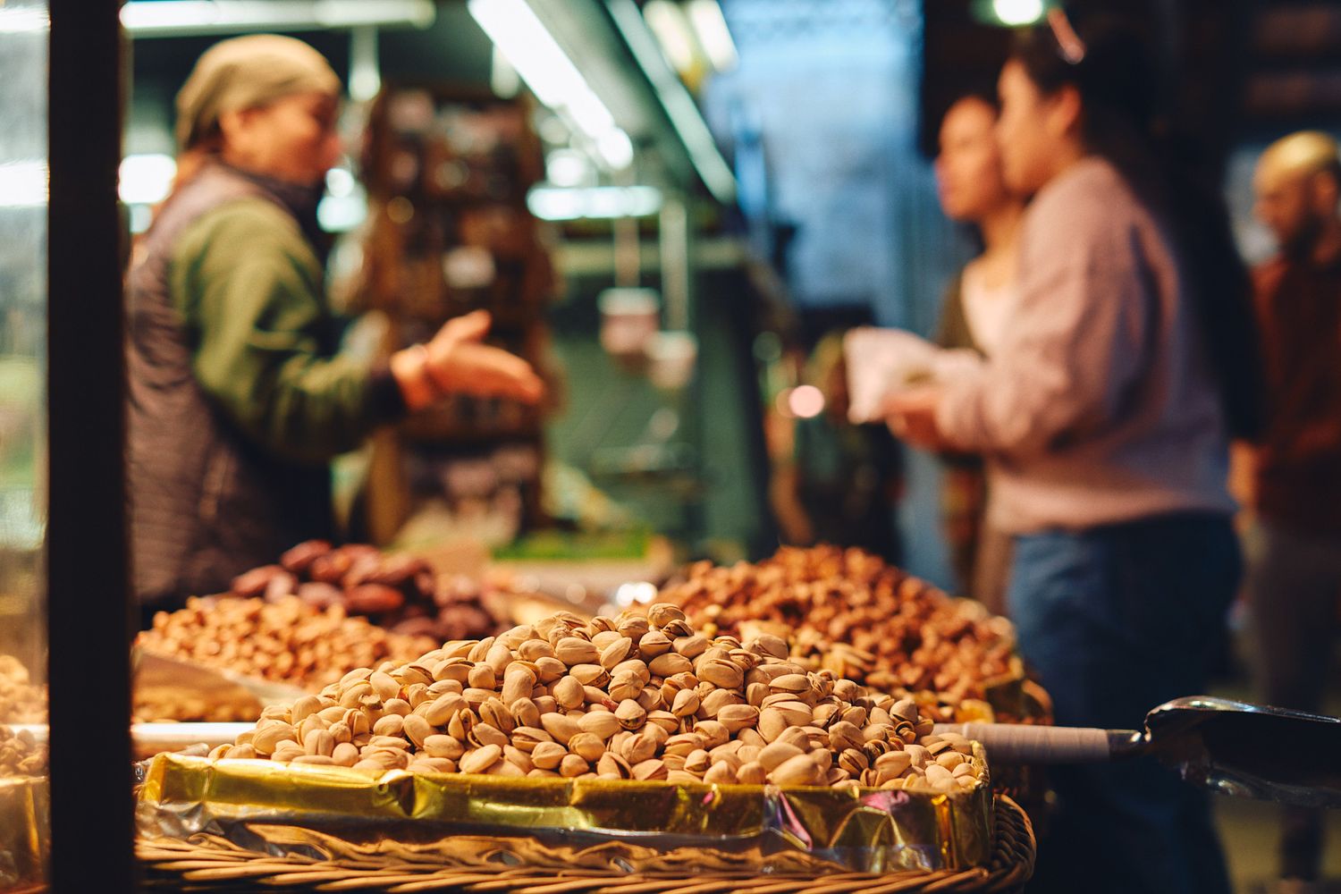 Nuts displayed in baskets at a bustling market stall with vendors and customers in the background.