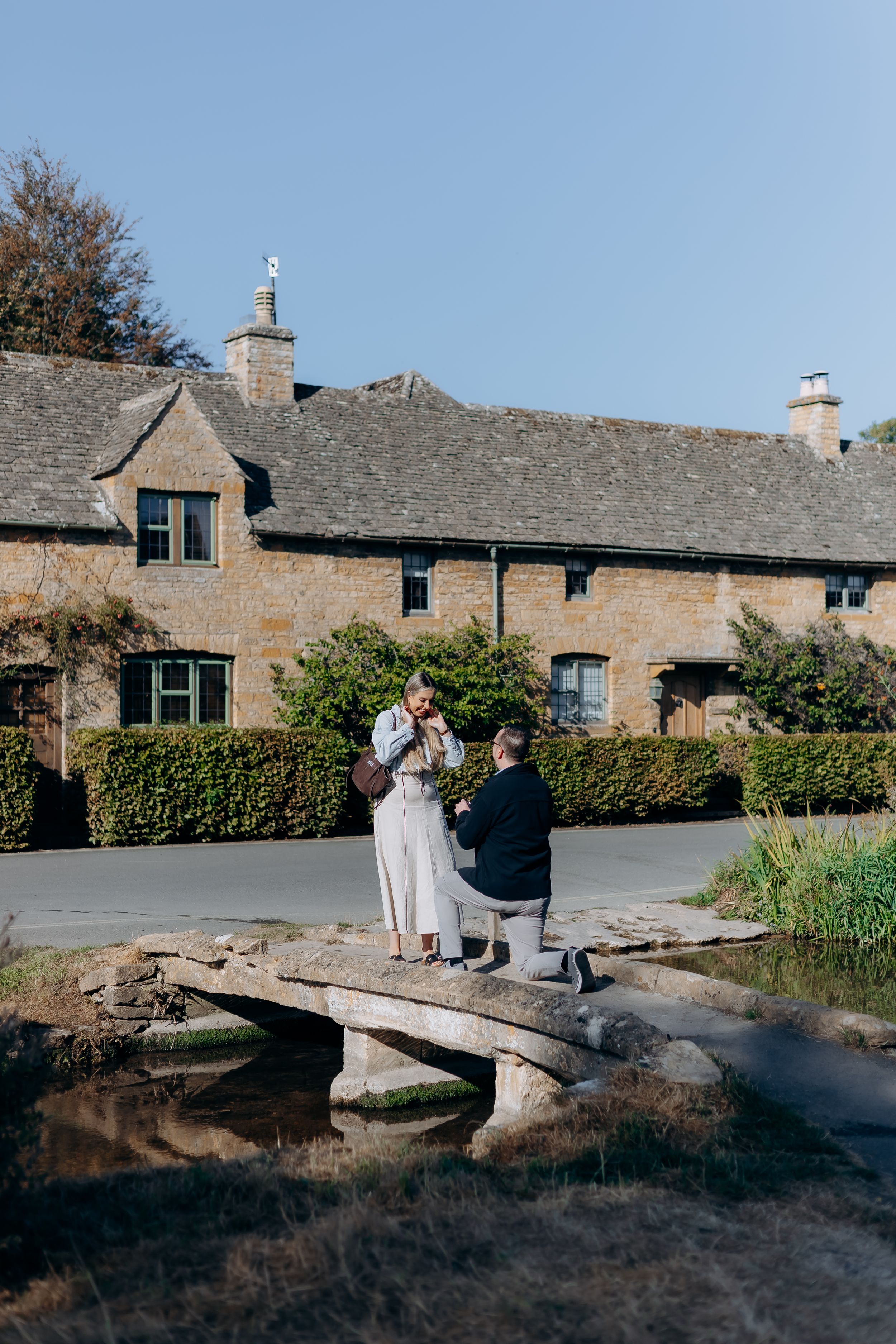 Man proposing to woman on stone bridge before charming Cotswolds village lower slaughter.