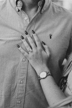 Close-up of a woman's hand with dark nails and diamond ring resting on a man's button-down shirt.