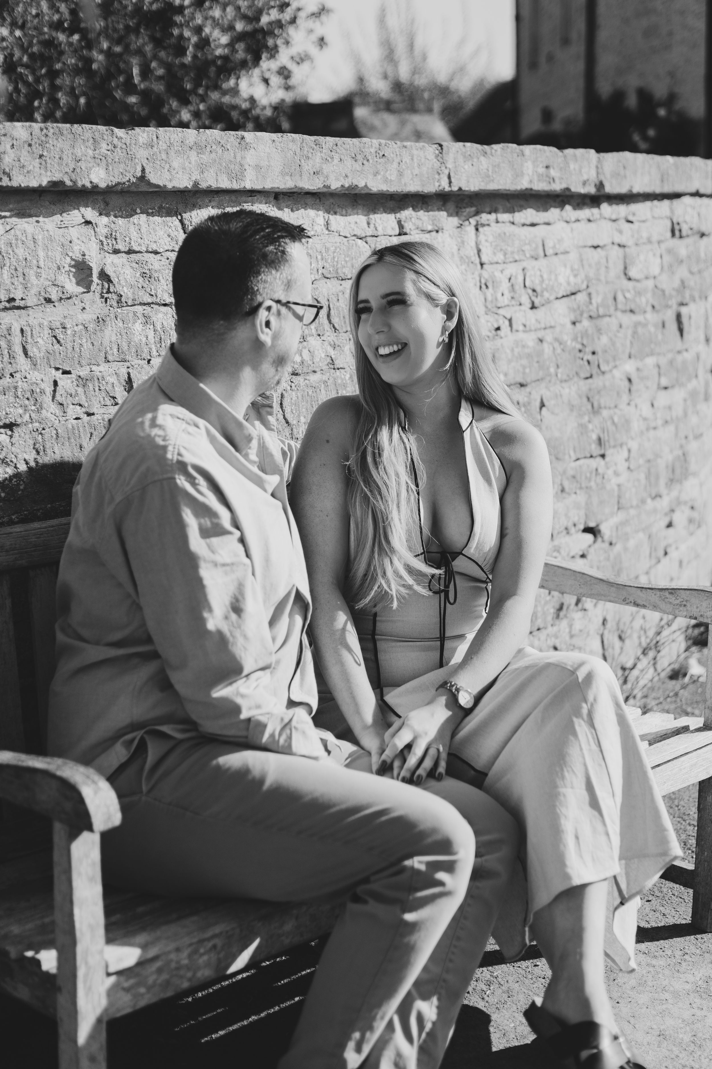 Black and white photo of a smiling couple sitting on a bench against a brick wall, enjoying a sunny day together in the Cotswolds.