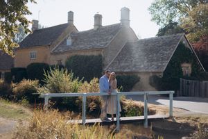Couple embracing on a white bridge beside a charming Cotswolds stone cottage surrounded by autumn foliage in Lower Slaughter.
