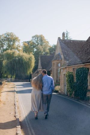 Couple walking arm in arm down a quiet village road lined with stone cottages and weeping willows on a sunny day in the Cotswolds.