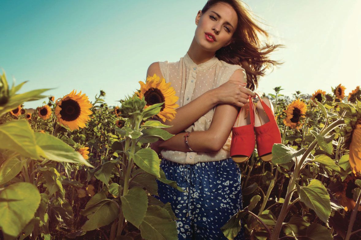 Woman in floral skirt holding red bag among sunflowers in sunny outdoor fashion shoot.