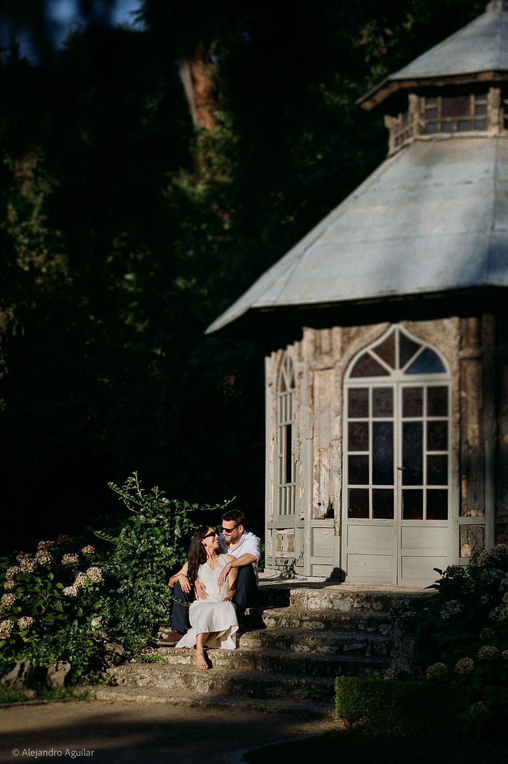 Couple sitting on steps of a charming stone pavilion surrounded by lush greenery in golden light.