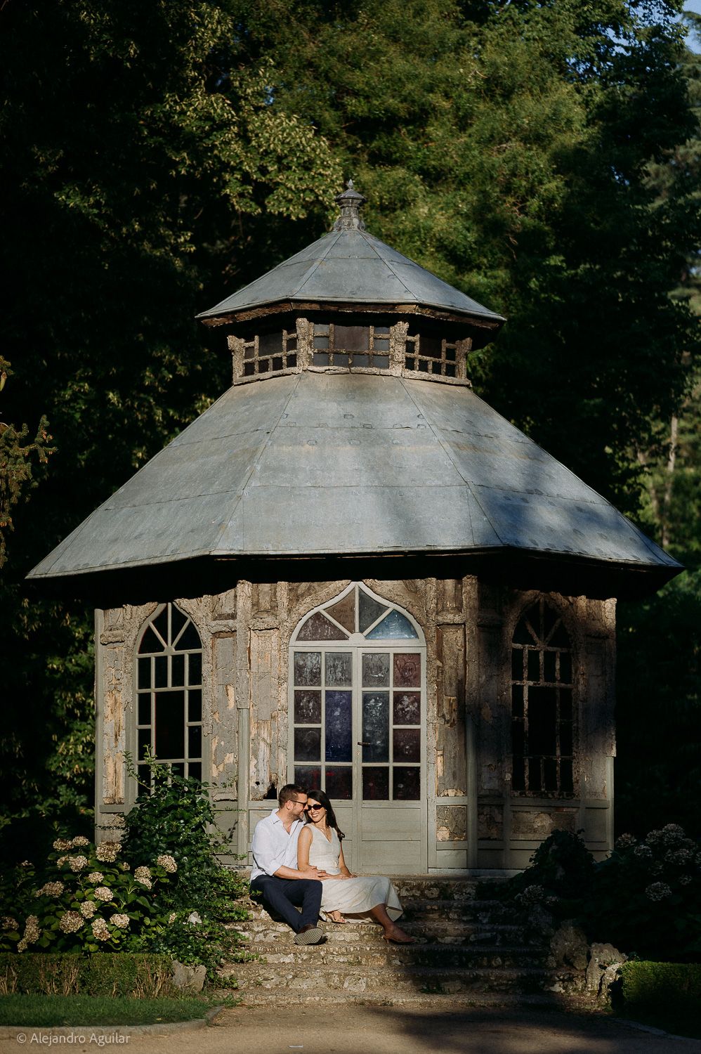 Couple sitting together at the entrance of a rustic stone gazebo surrounded by lush green trees and hydrangeas.