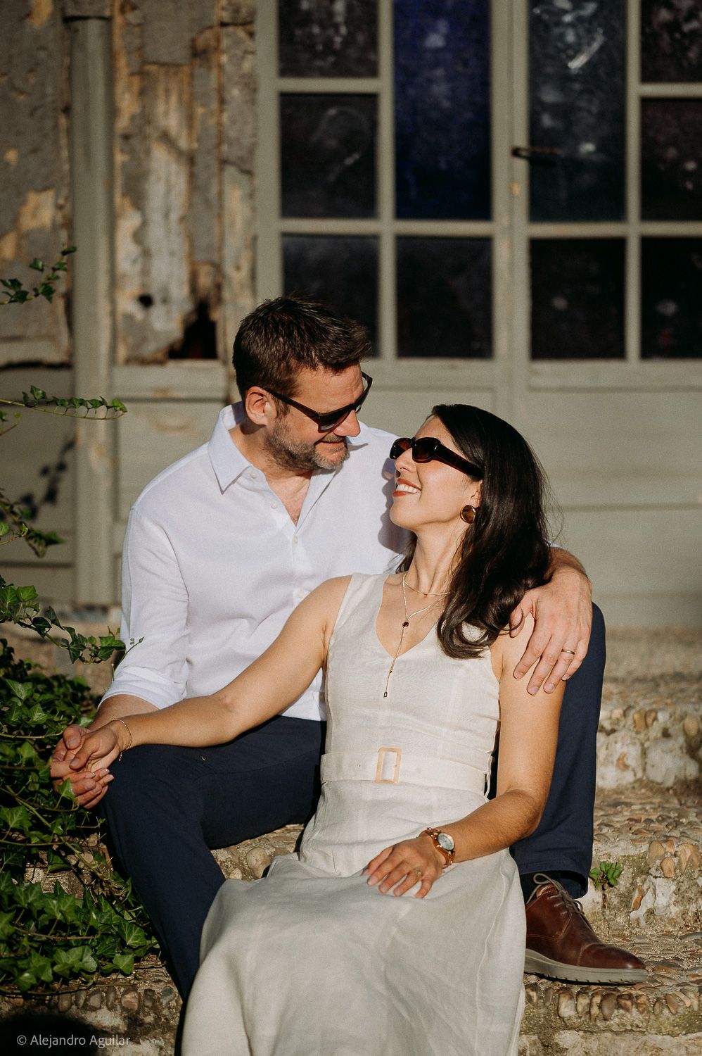 Couple sitting together outdoors, smiling and wearing sunglasses, in front of a rustic building.