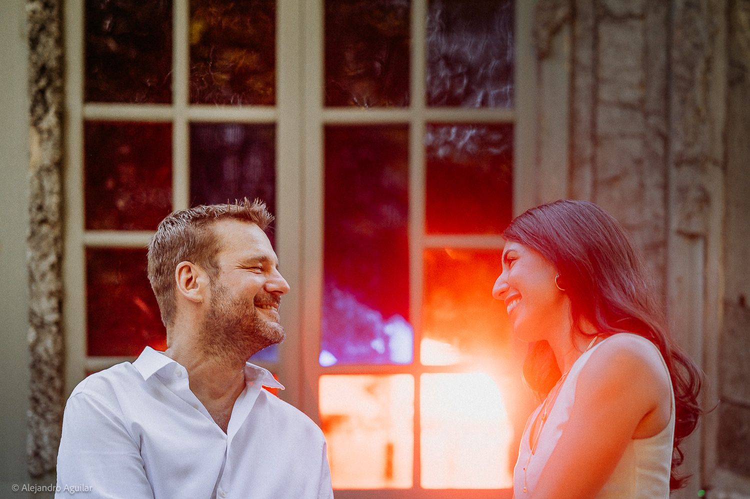 A smiling couple shares a joyful moment outdoors with warm sunlight glowing through French doors behind them.
