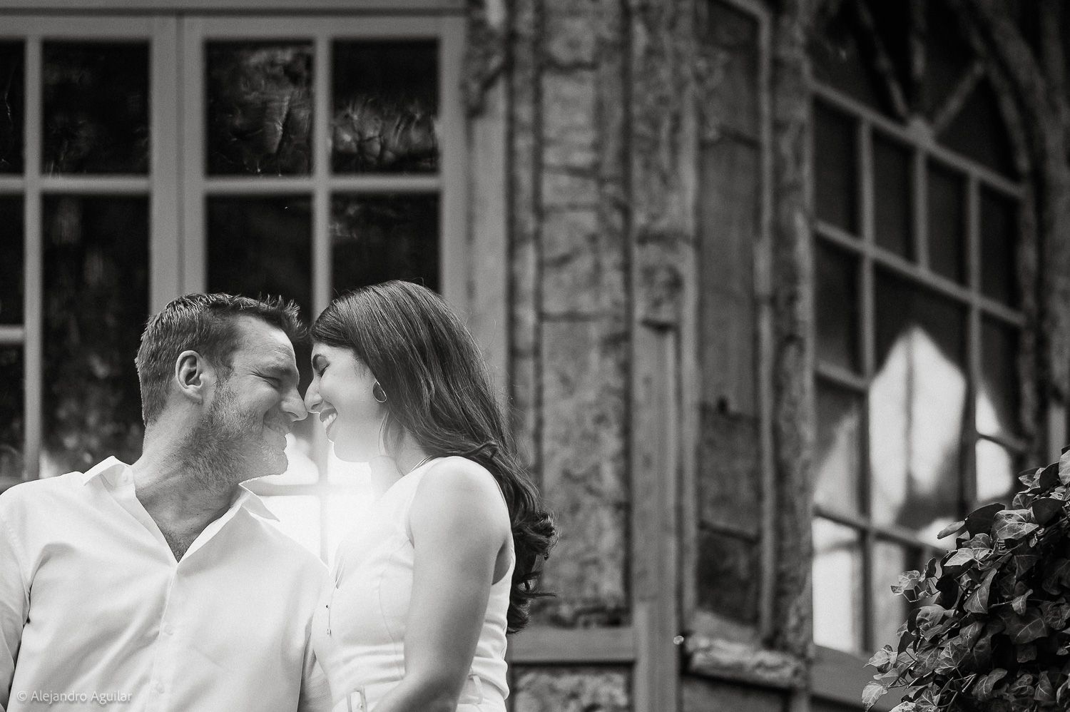 A romantic couple shares an intimate moment in black and white, leaning close together near rustic stone windows.