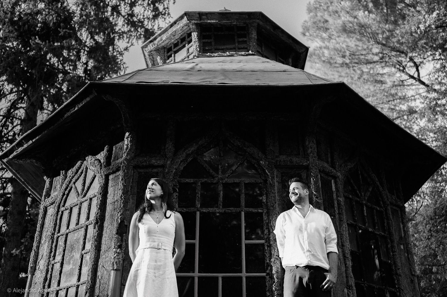 Black and white photo of a couple standing before a rustic wooden gazebo surrounded by trees.
