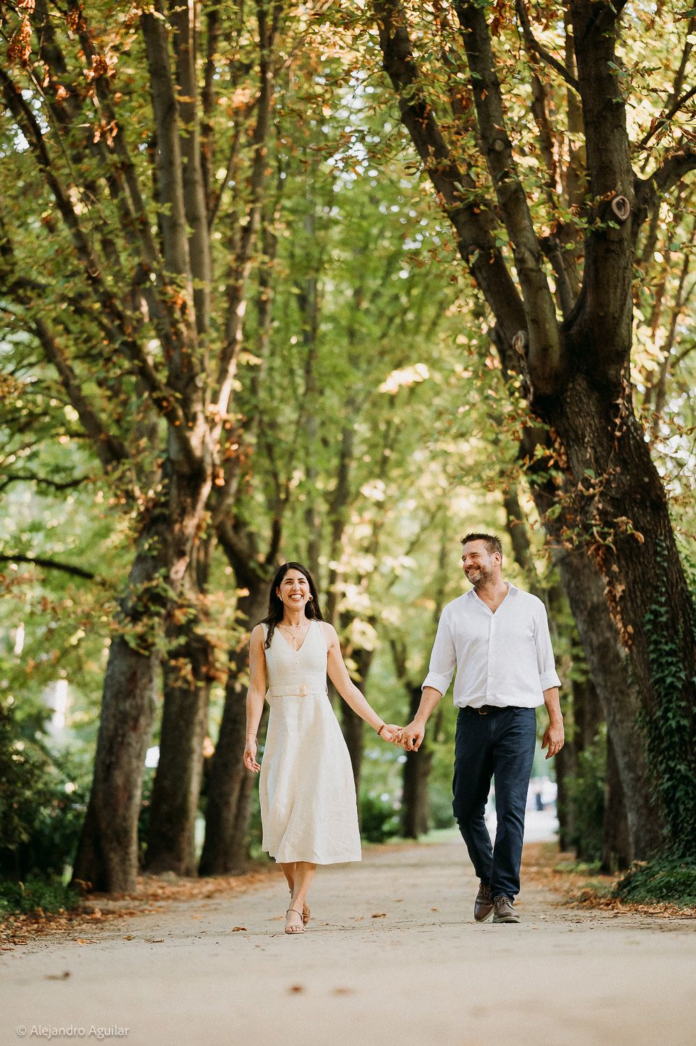 A couple walks hand-in-hand down a tree-lined path, smiling, woman in white dress, man in white shirt and dark pants.