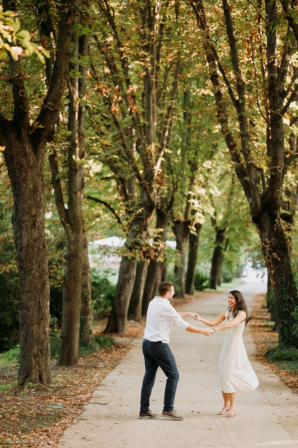 A couple dances joyfully on a tree-lined path surrounded by lush autumn foliage in a romantic outdoor setting.
