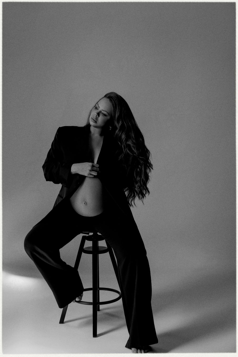 Black and white maternity photo of a woman in a suit sitting on a stool, showing her baby bump in a studio.