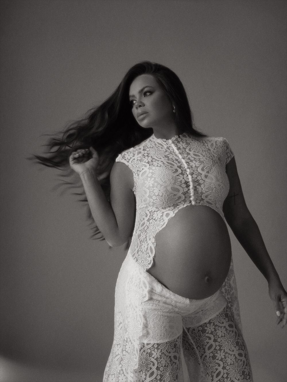 Black and white maternity photo of a pregnant woman in white lace dress with flowing hair in a studio.