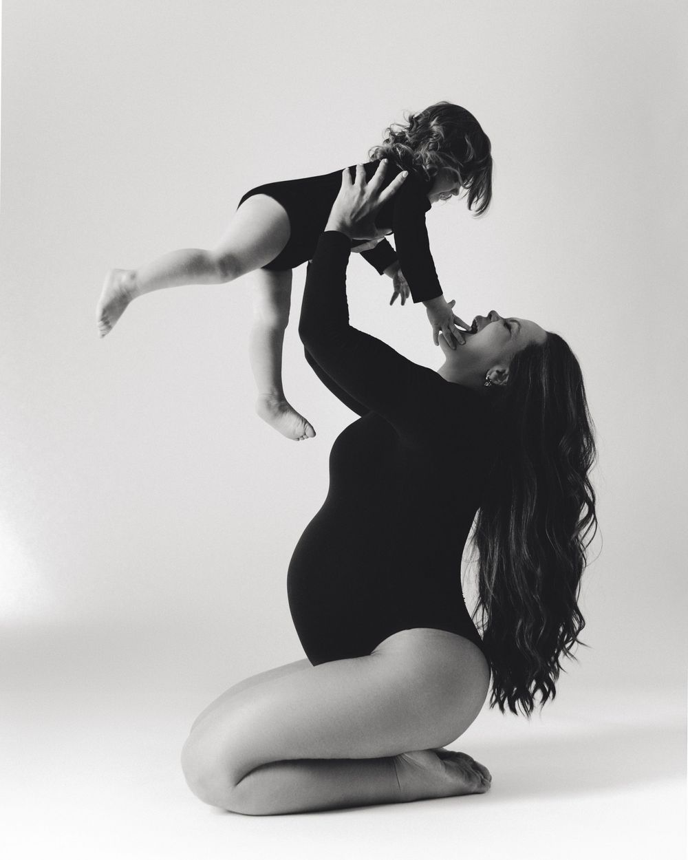 Black and white photo of a mother kneeling and lifting her baby overhead, both wearing black outfits.