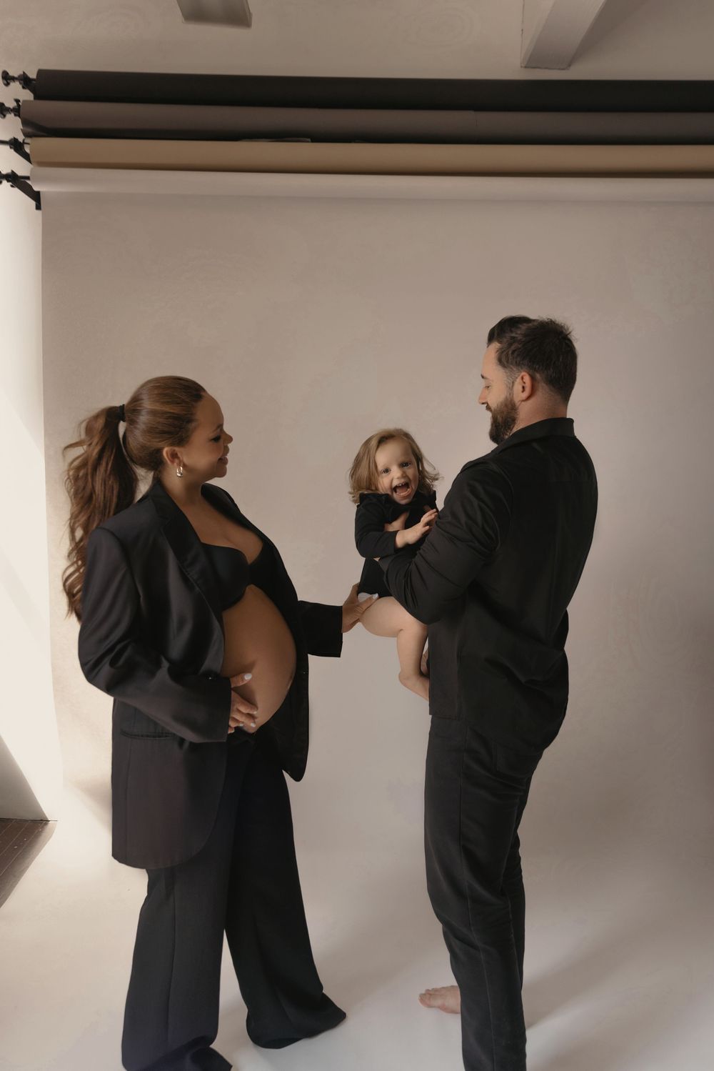 Pregnant woman in black suit with husband holding toddler, family maternity photo session in studio.
