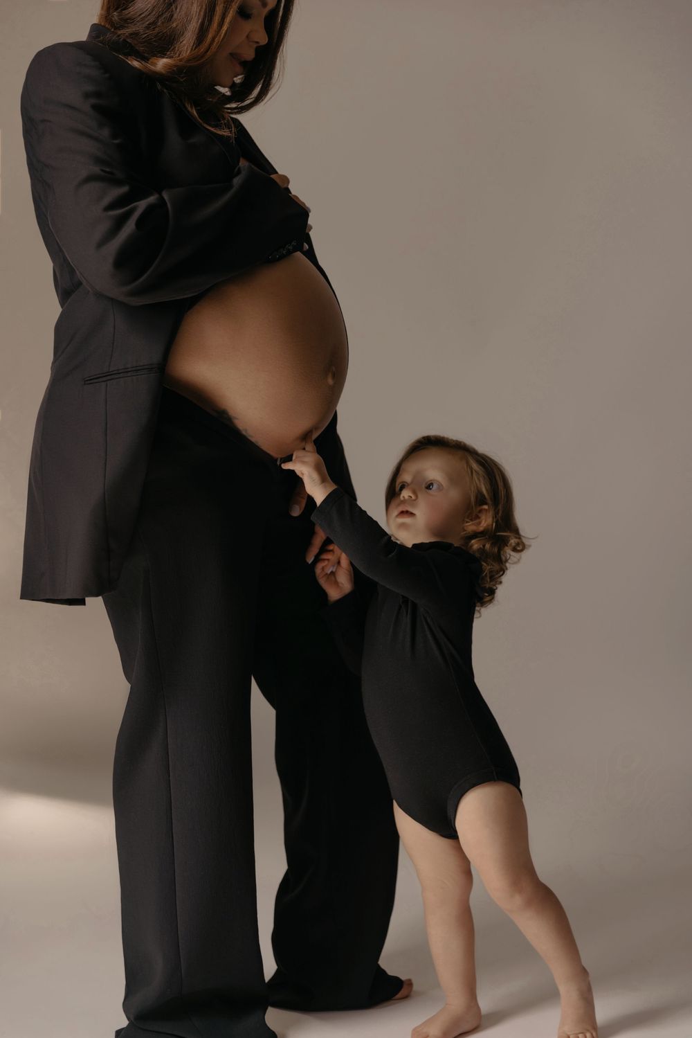 Pregnant woman in black suit poses with toddler in black bodysuit touching her baby bump in studio.