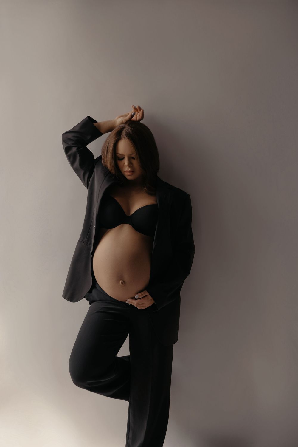 Pregnant woman in black blazer and bra cradling baby bump against gray wall.