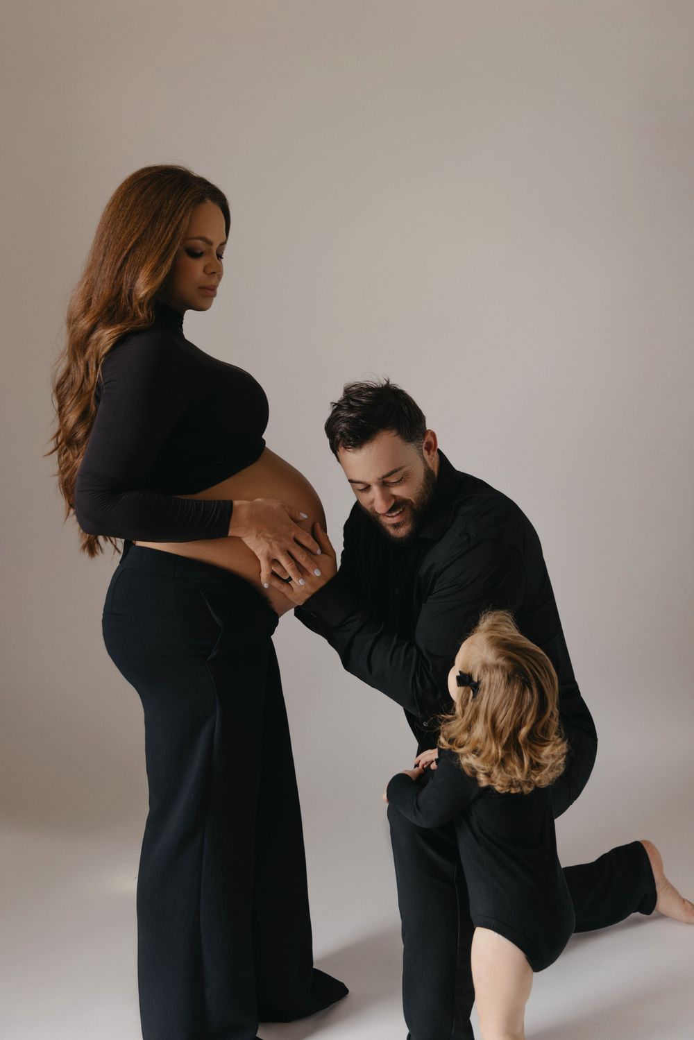 A pregnant woman in black with her partner and child touching her baby bump during a maternity photoshoot.