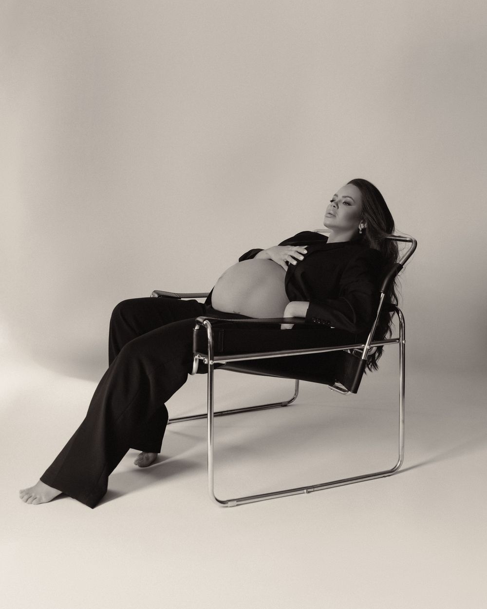 Black and white maternity photo of a pregnant woman reclining in a modern chrome chair, wearing a black outfit.