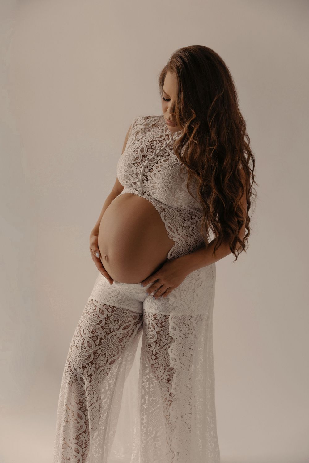 Pregnant woman in white lace dress cradling baby bump, long wavy hair, elegant maternity photo on light background.