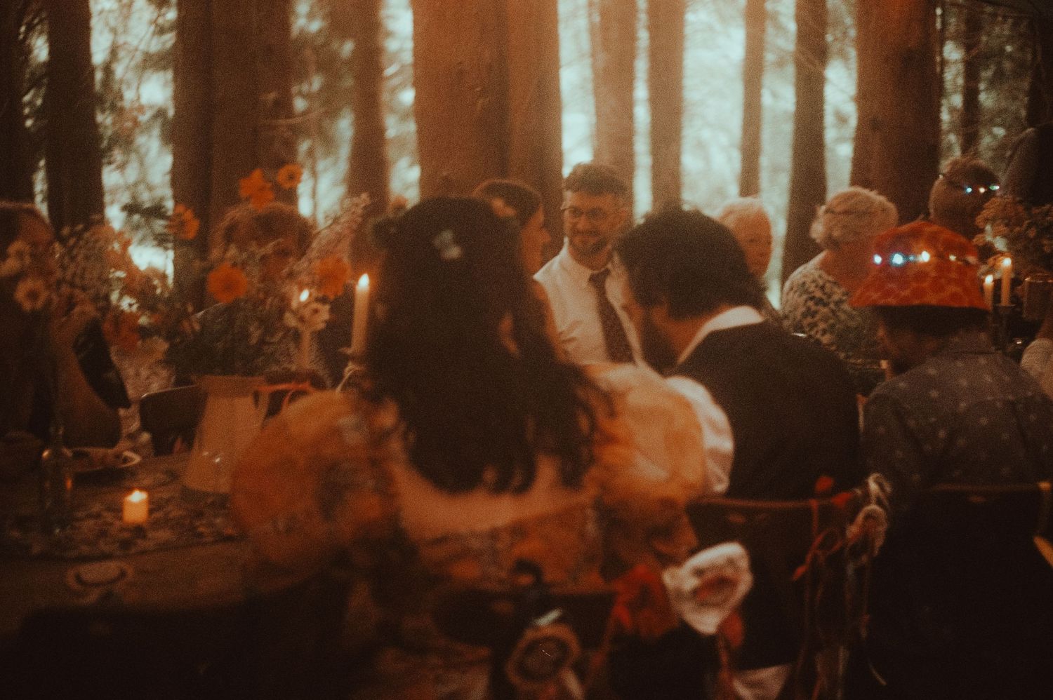 Romantic outdoor wedding reception dinner in a forest setting with warm candlelight and guests gathered around a decorated table.