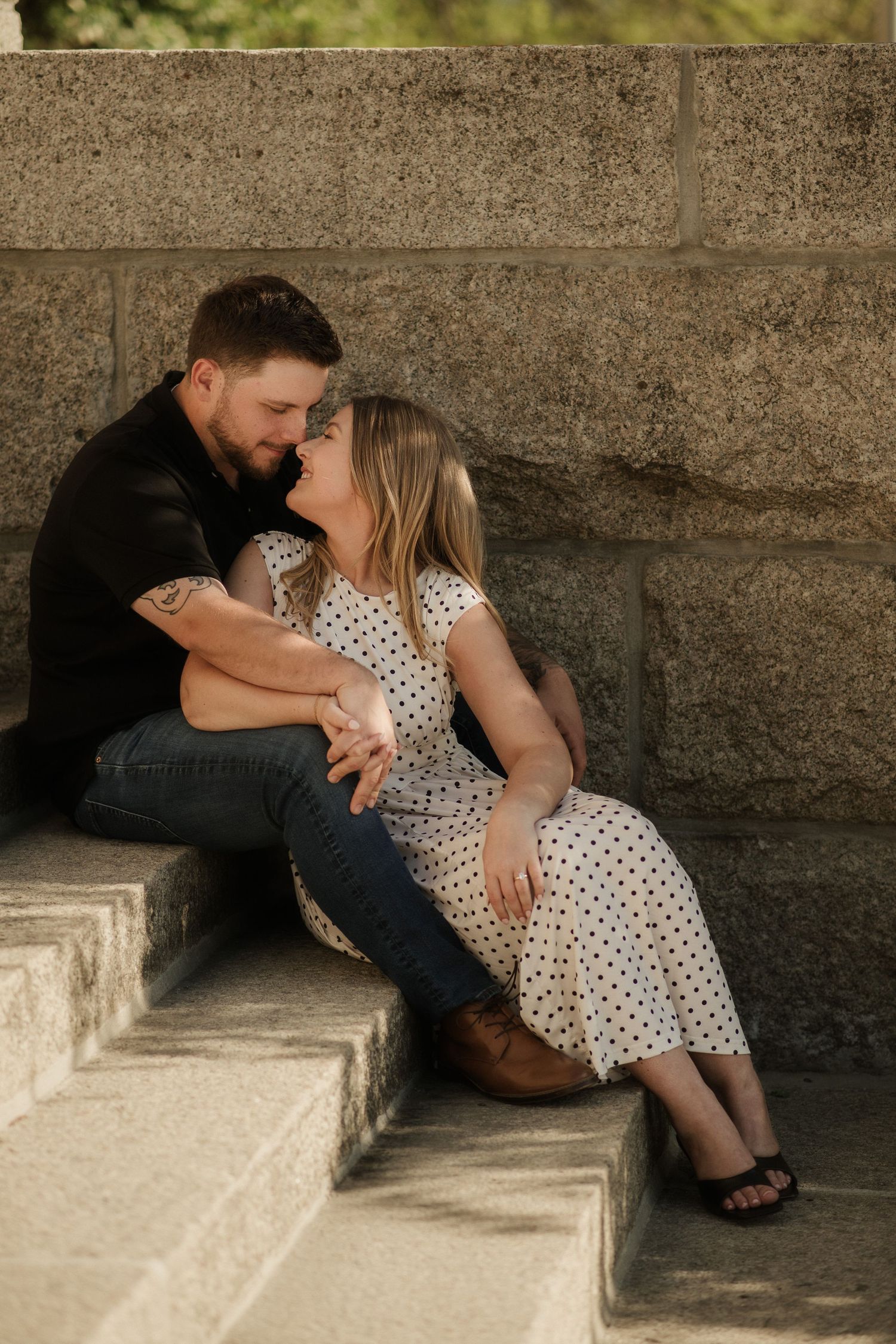 Couple sharing a romantic moment on stone steps, woman in polka dot dress leaning toward man in black shirt.