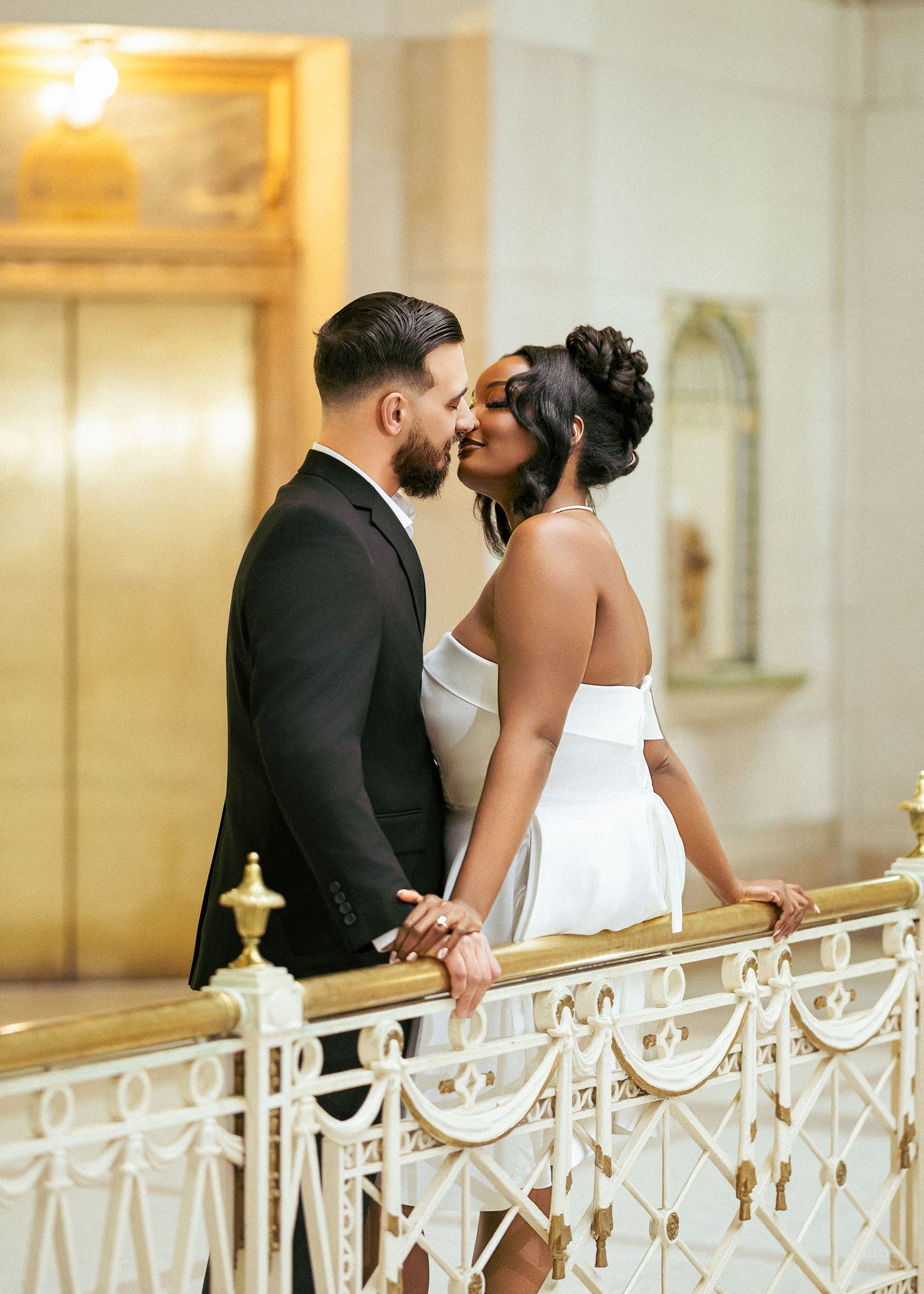 Couple sharing a romantic almost-kiss on an ornate gold railing staircase, bride in white dress and groom in black suit.