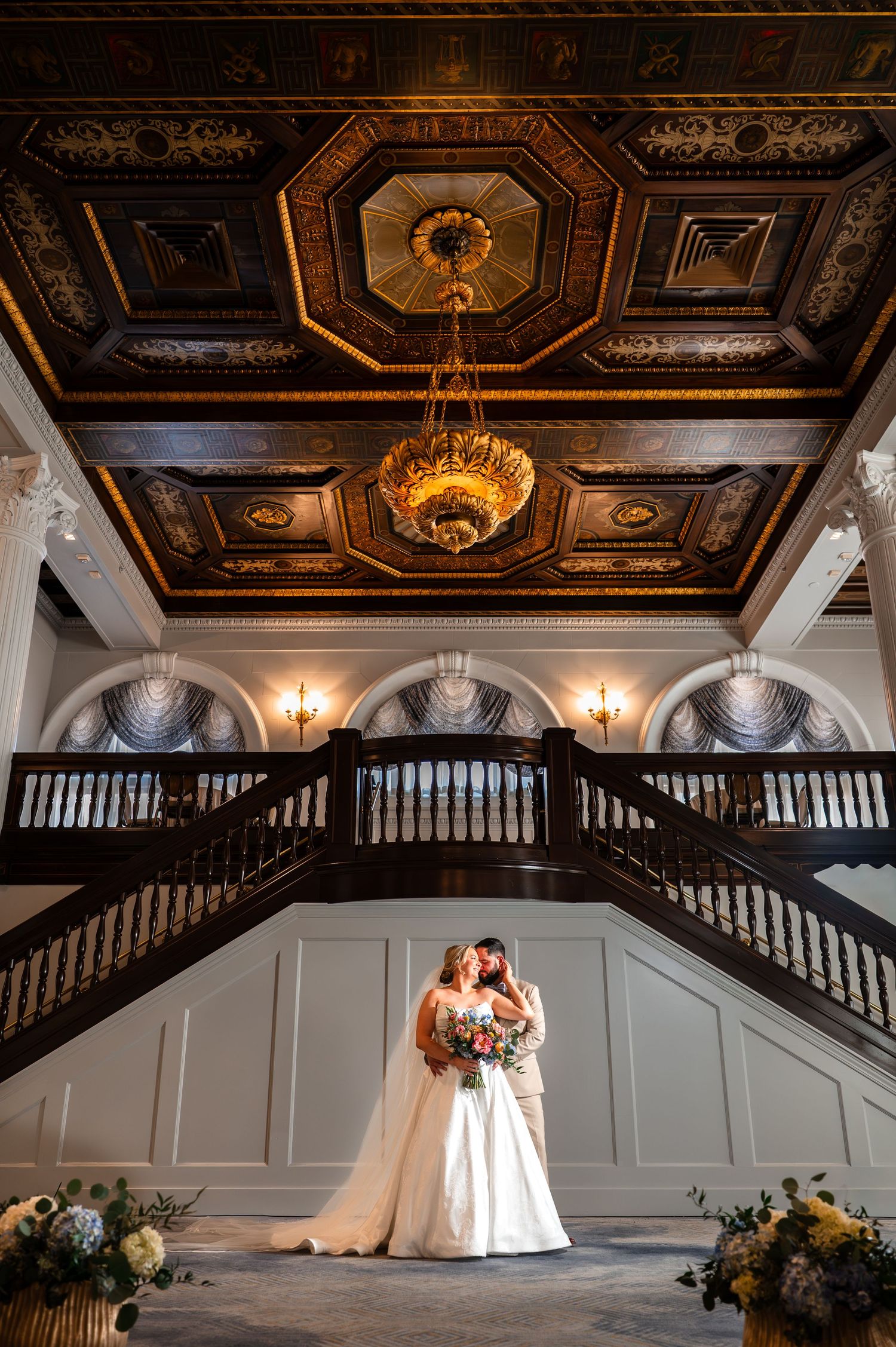 Bride and groom embrace under ornate coffered ceiling with gold chandelier on grand staircase in elegant wedding venue.