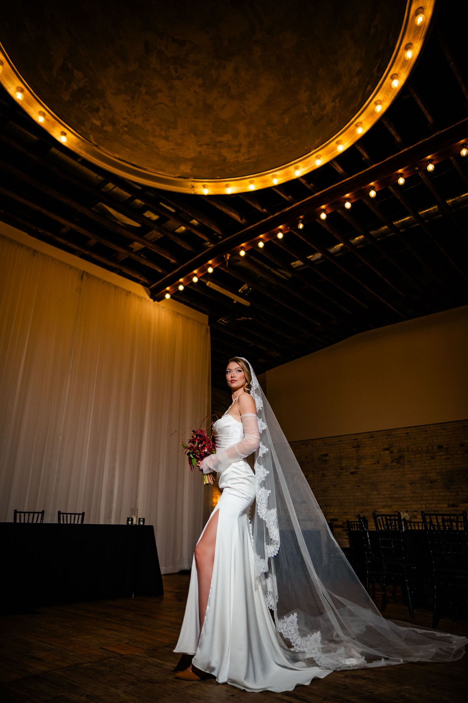 Bride in white gown with long veil holds bouquet in elegant venue with marquee lights and draped curtains.
