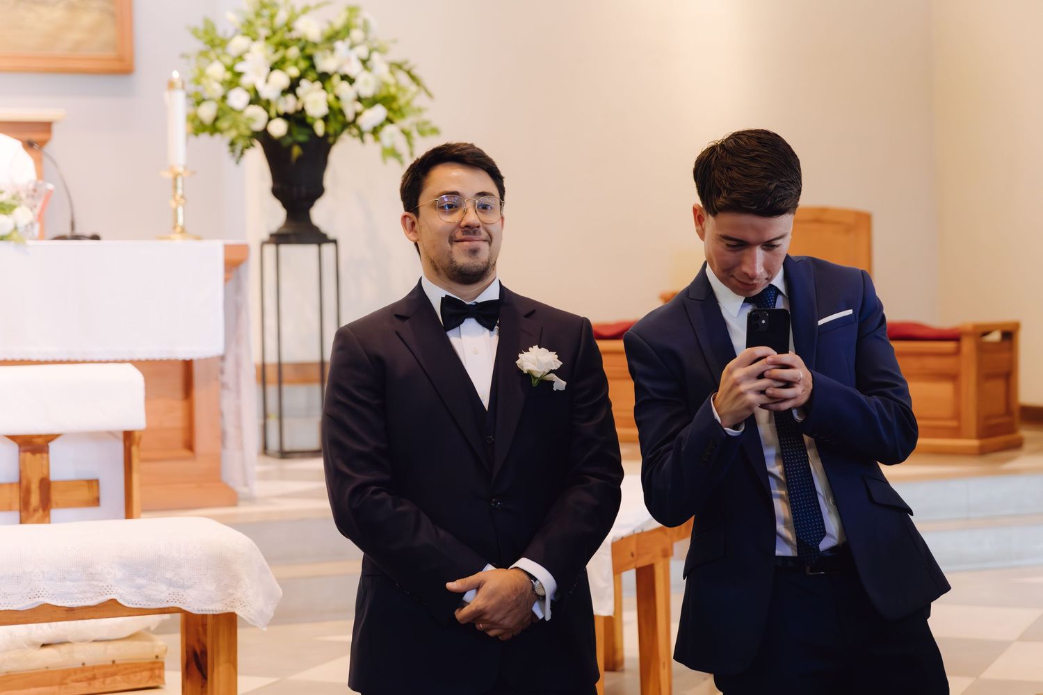Groom in dark tuxedo with boutonniere standing at church altar with groomsman before wedding ceremony.