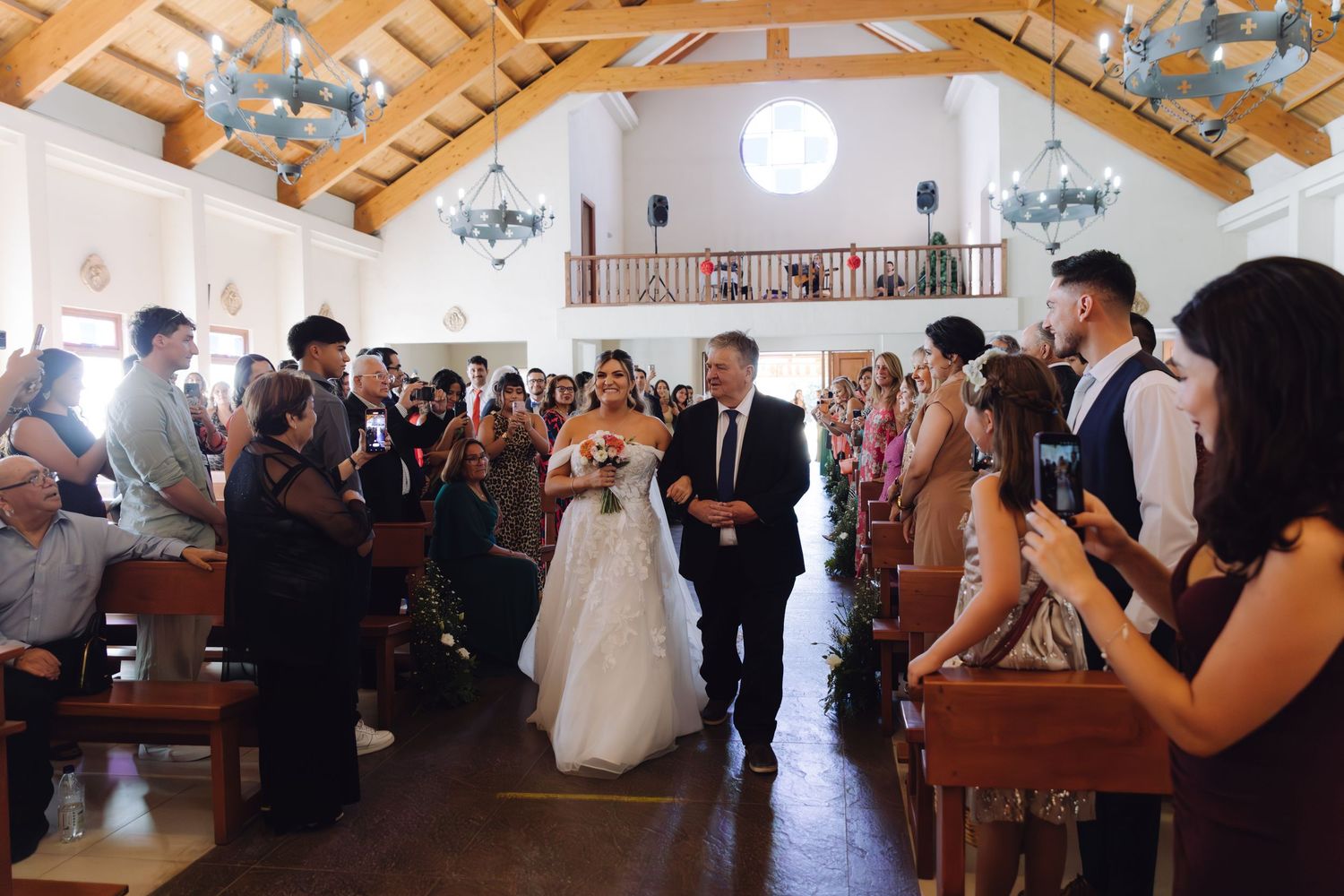 Bride in white gown walking down church aisle with escort as guests photograph the wedding processional.