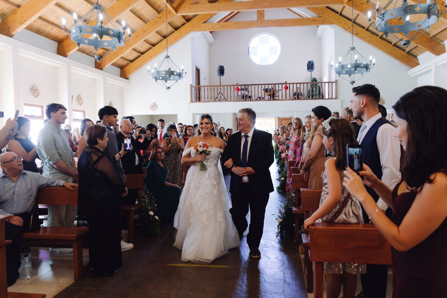 Elegant bride carrying bouquet walks down decorated church aisle with guests capturing the wedding processional.