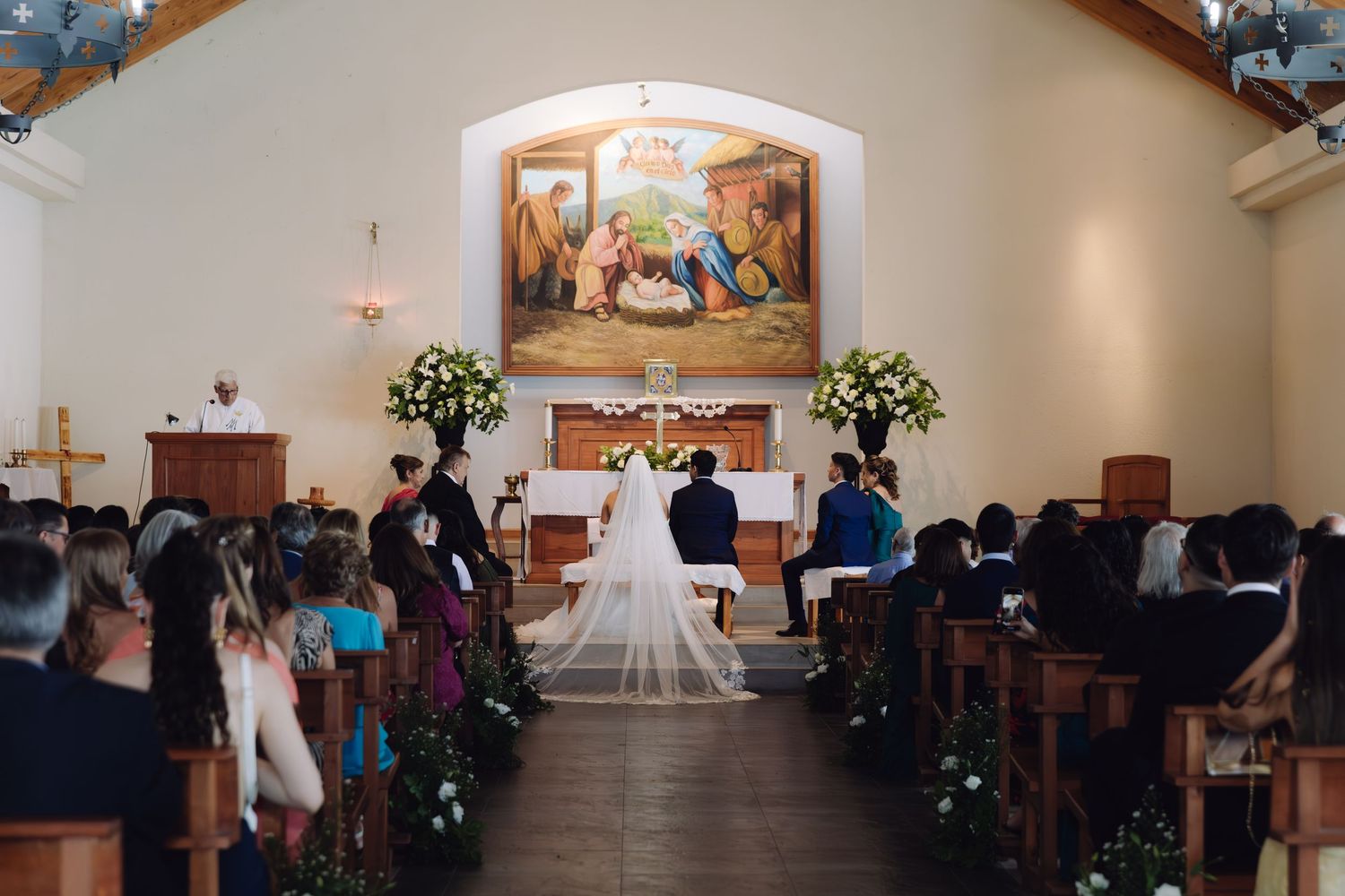 Wide view of traditional Catholic church wedding ceremony with bride and groom at ornate floral altar.