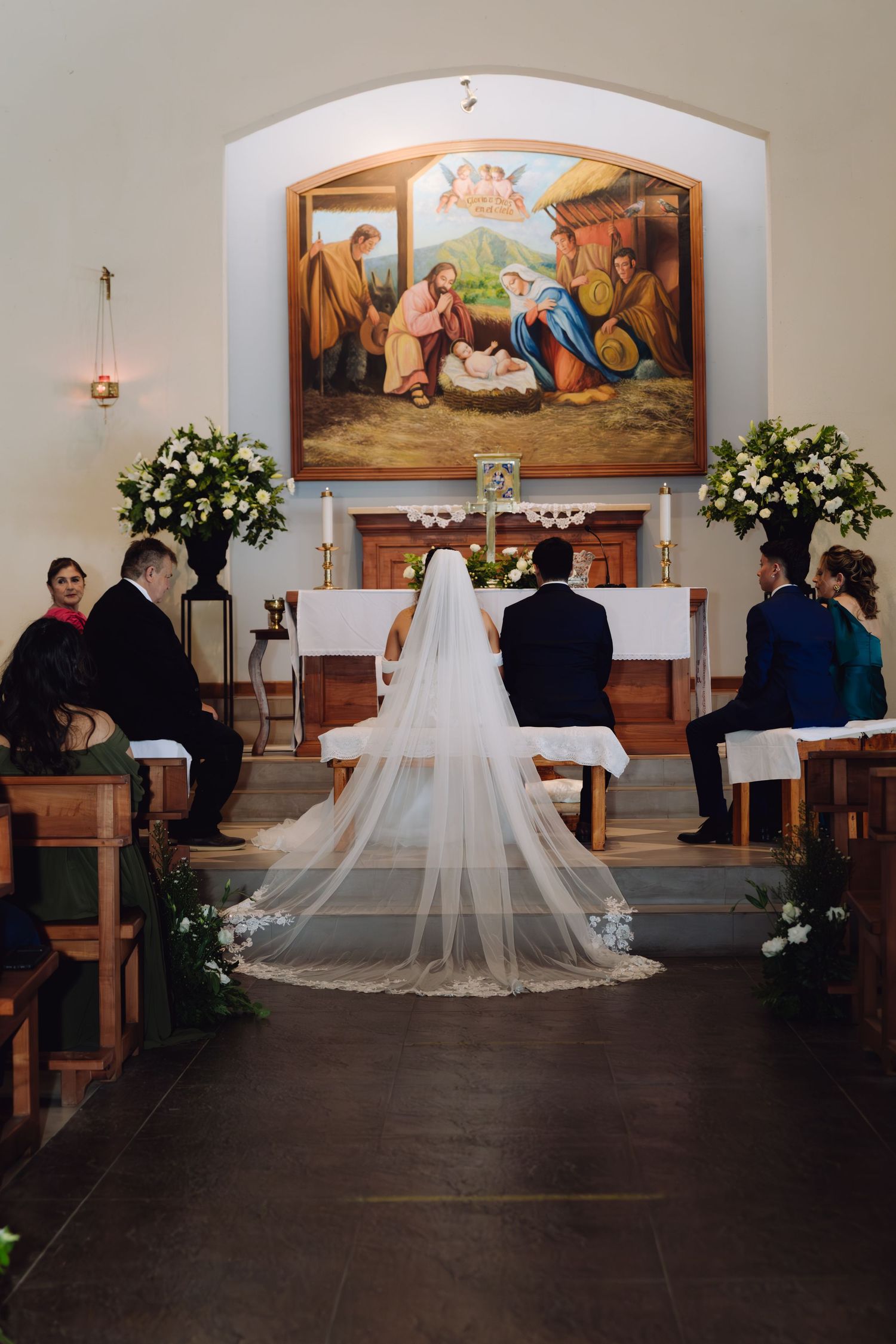 Bride in long cathedral veil kneeling with groom before nativity painting at Catholic wedding ceremony.