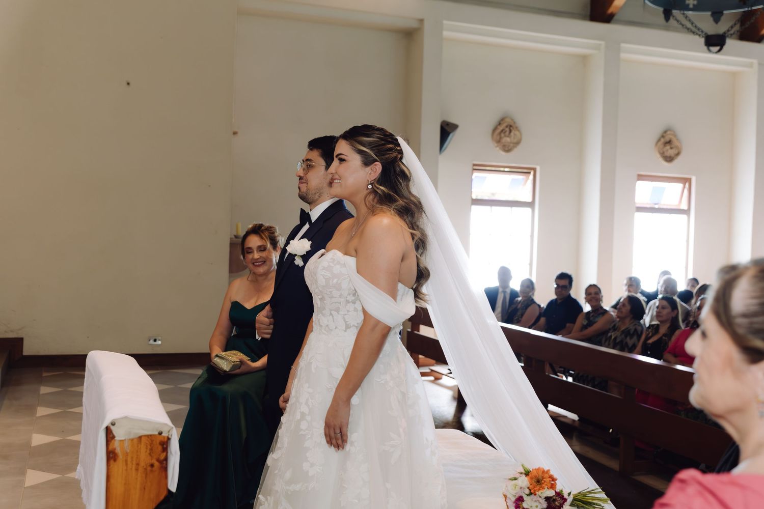 Bride in off-shoulder gown and long veil standing with groom during church wedding ceremony.