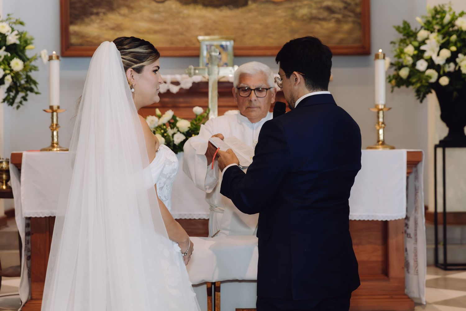 Bride and groom exchanging rings before elderly priest at decorated church altar during wedding ceremony.