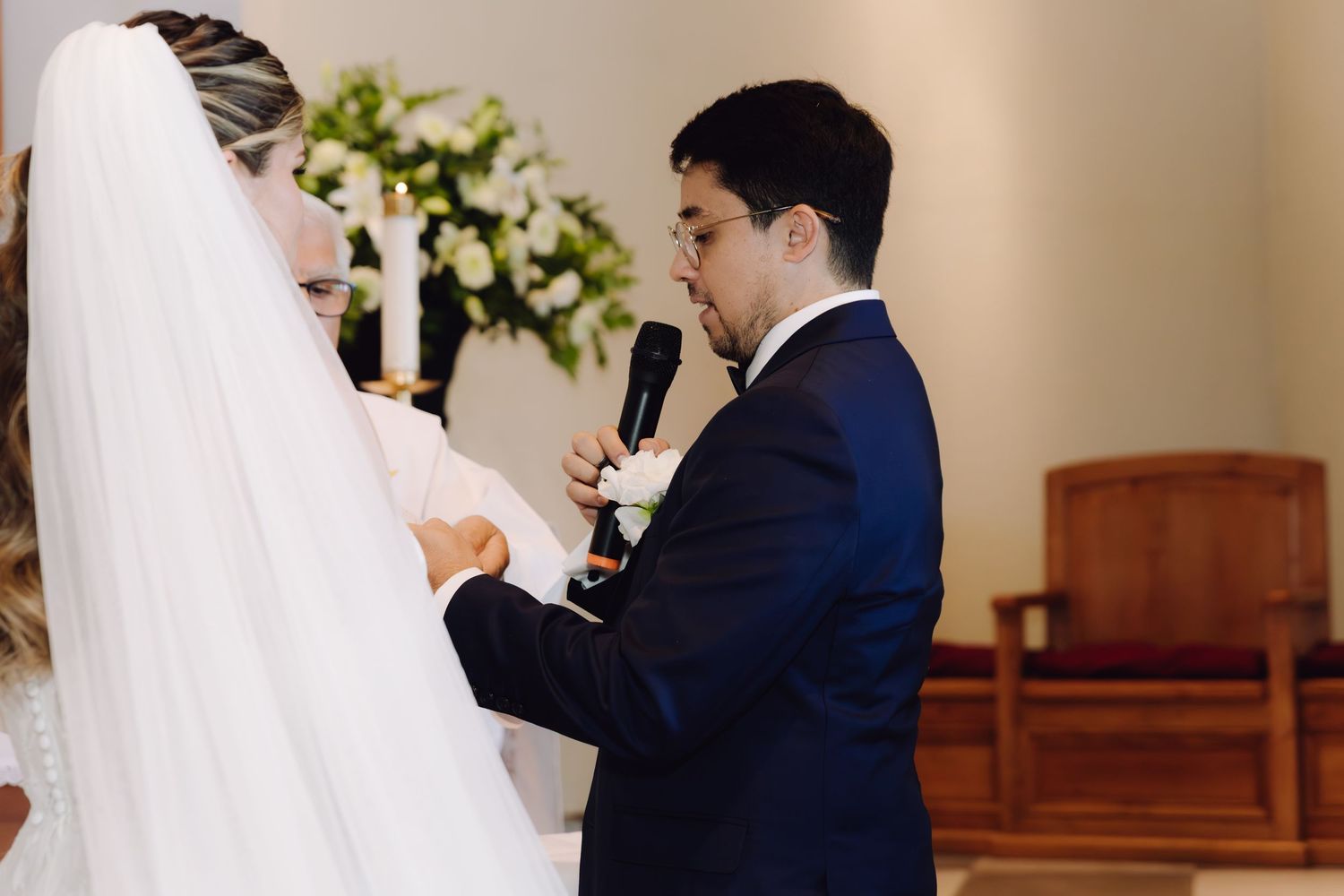 Groom in navy suit reciting wedding vows into microphone while holding brides hands at church altar.