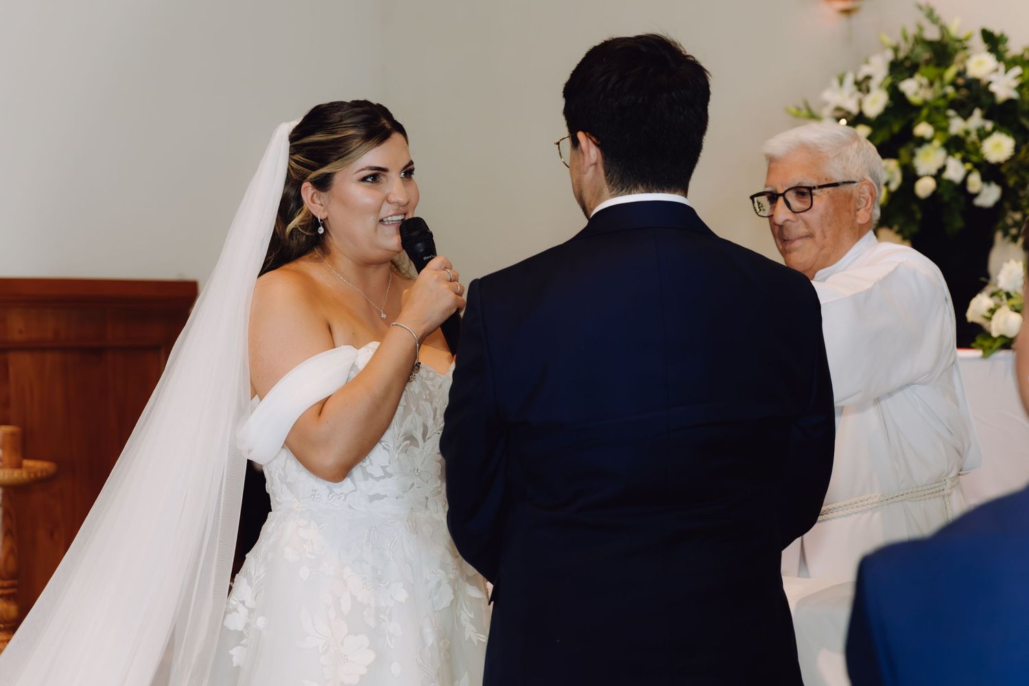 Smiling bride in white gown reciting vows to groom as elderly officiant watches during church ceremony.