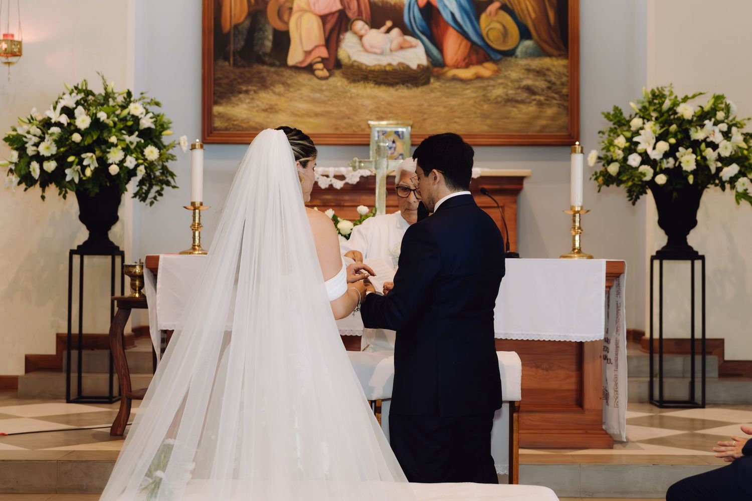 Bride and groom exchanging rings at church altar adorned with white floral arrangements and religious painting.