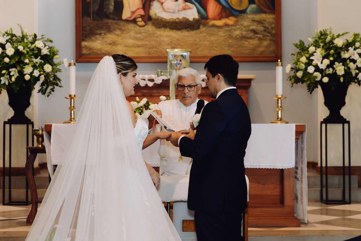 Elderly priest officiating ring exchange between bride in white gown and groom in navy suit at church altar.