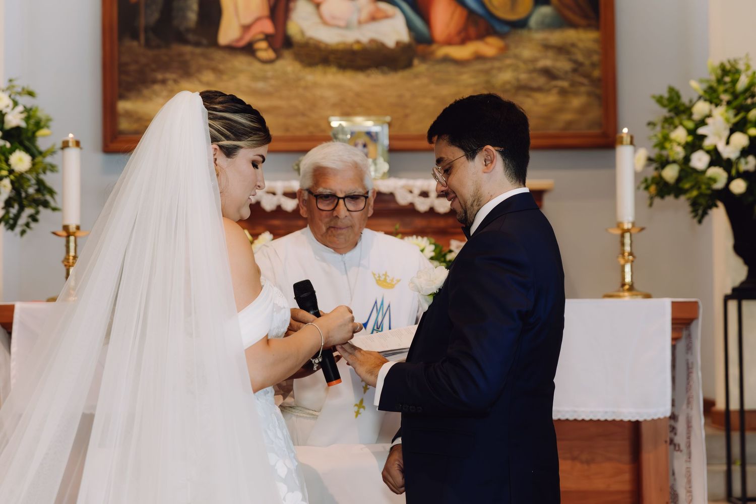 Bride placing wedding ring on grooms finger during Catholic church ceremony with priest officiating.