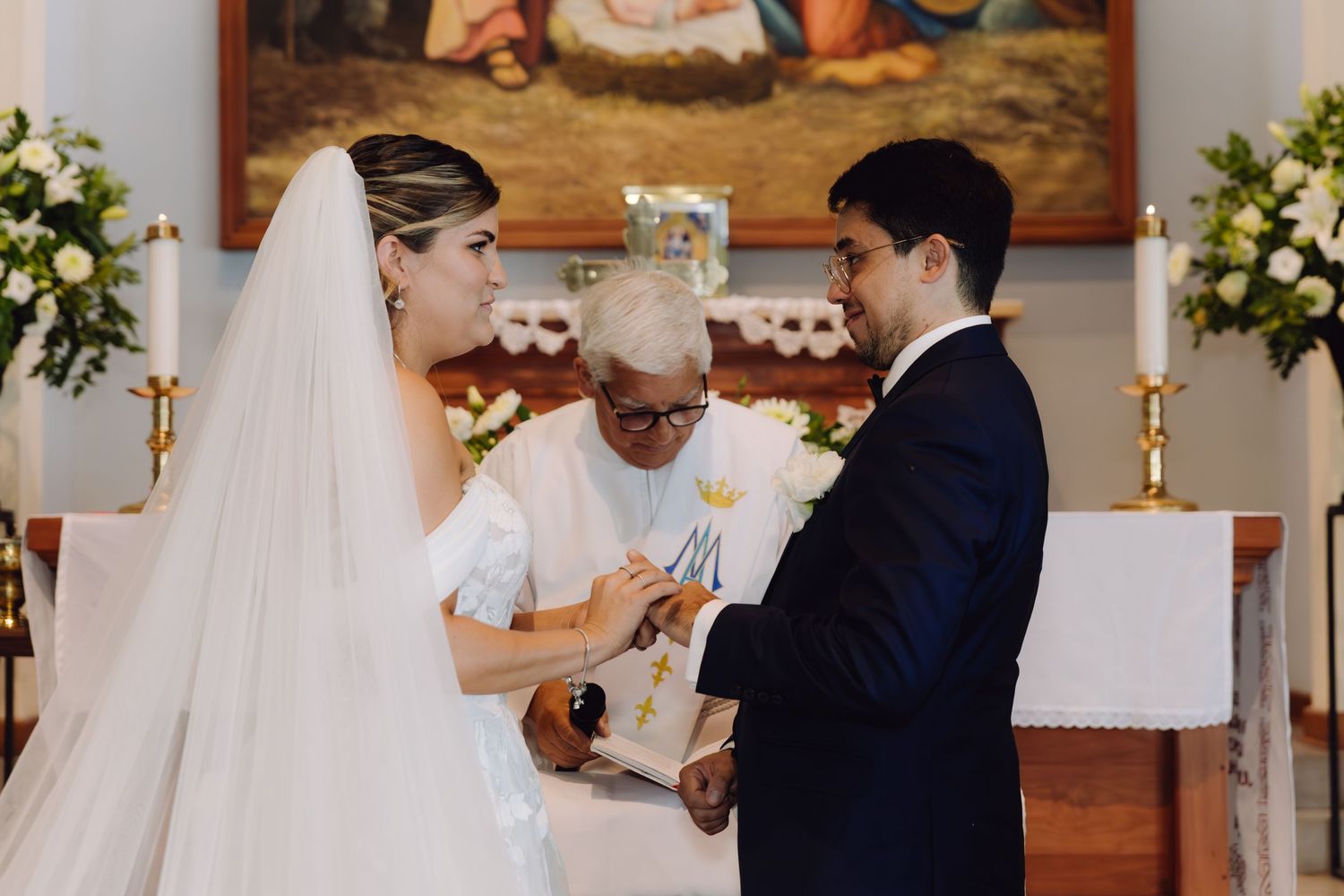 Smiling bride and groom exchanging rings with priest during elegant Catholic wedding ceremony at church altar.