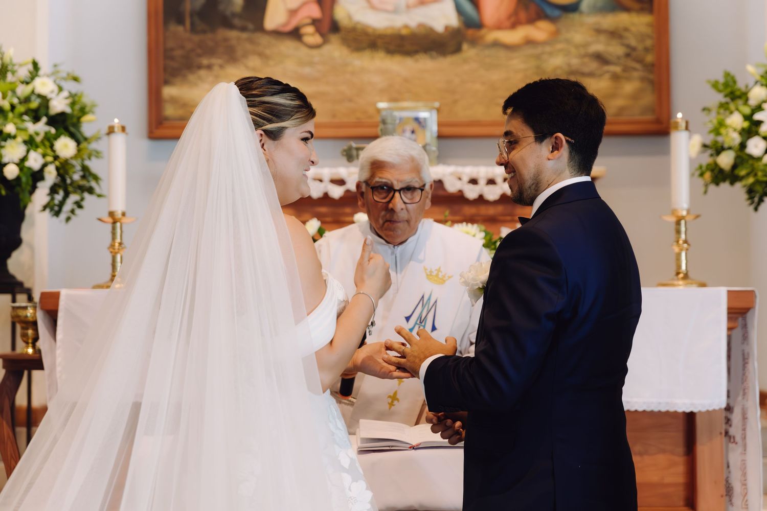 Bride and groom holding hands during ring exchange with elderly priest at decorated Catholic church altar.