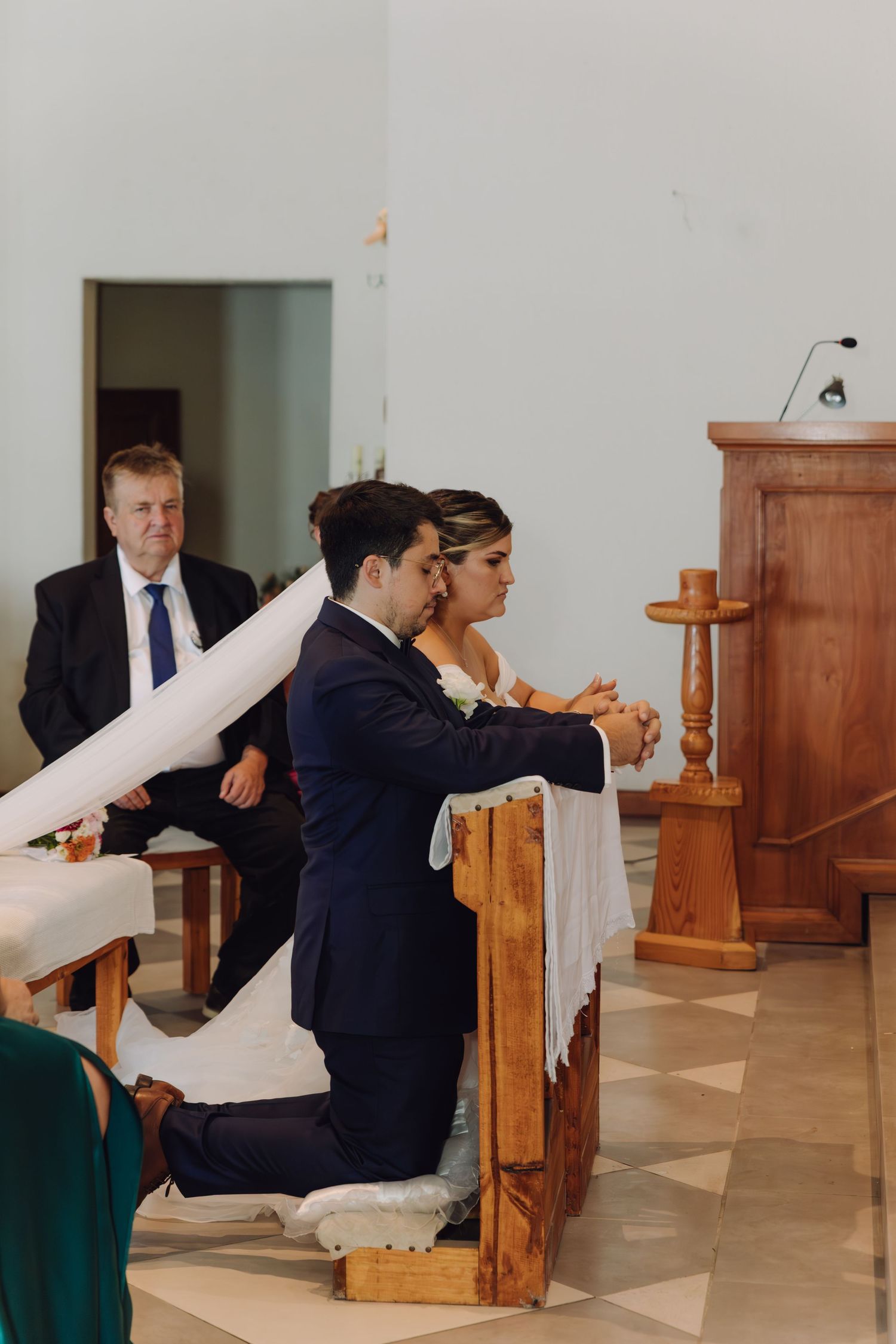 Bride and groom kneeling in prayer at wooden kneeler during Catholic wedding ceremony inside church.