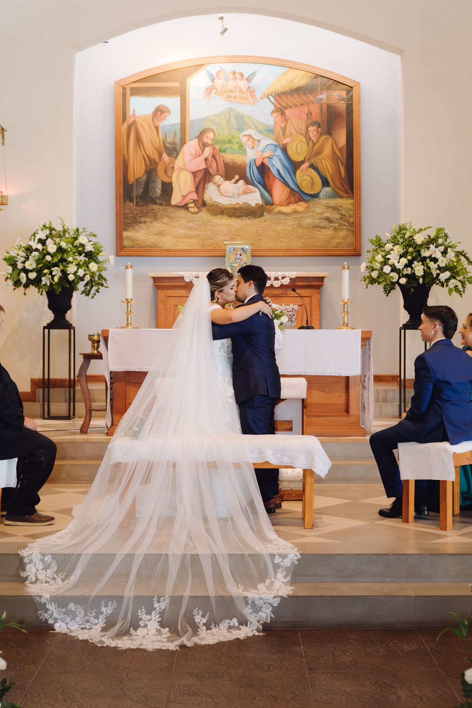 Bride and groom sharing first kiss at altar beneath nativity painting with lace cathedral veil trailing.