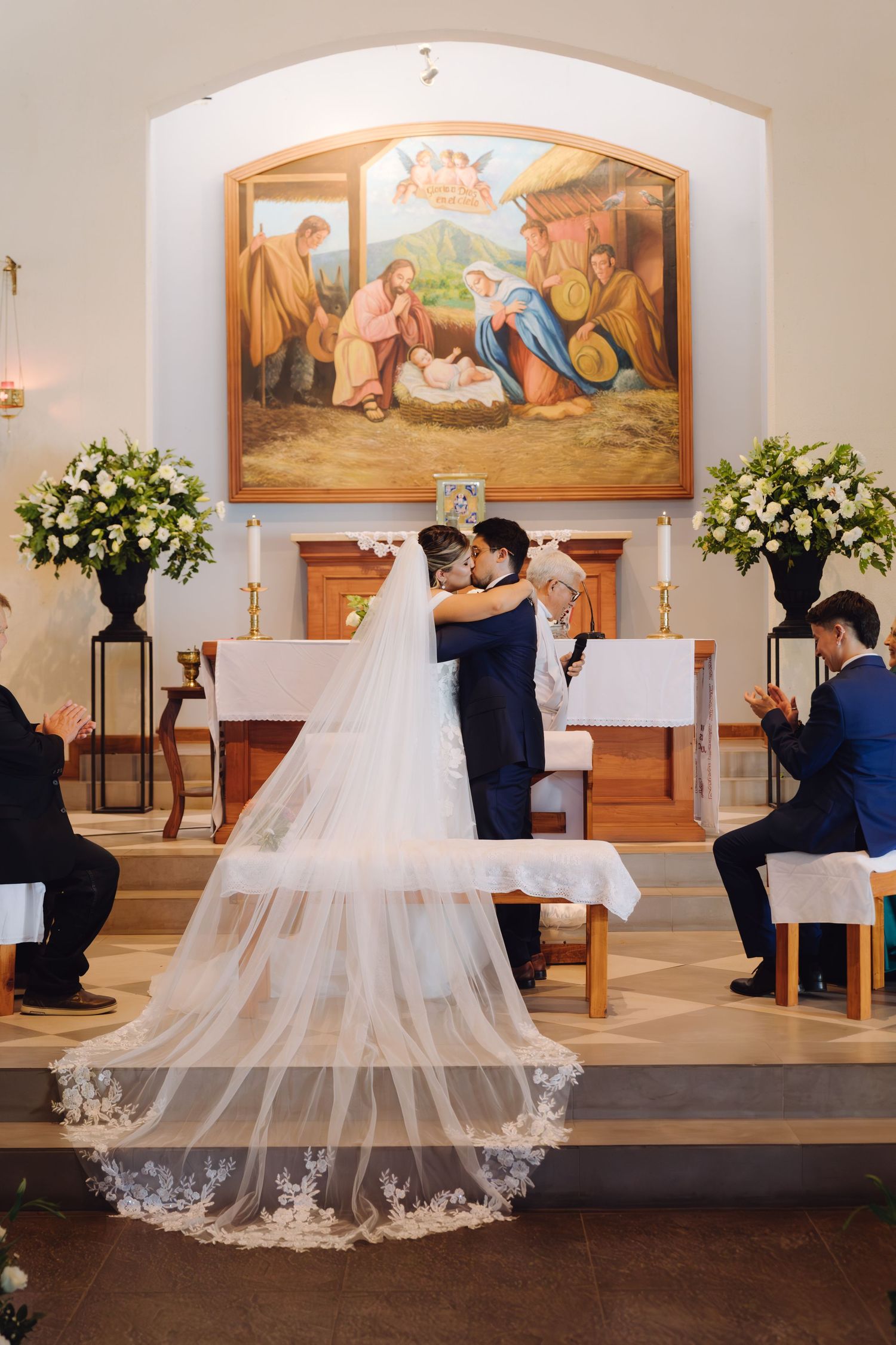 Newlyweds embracing at church altar as guests applaud beneath a nativity mural with white floral decor.