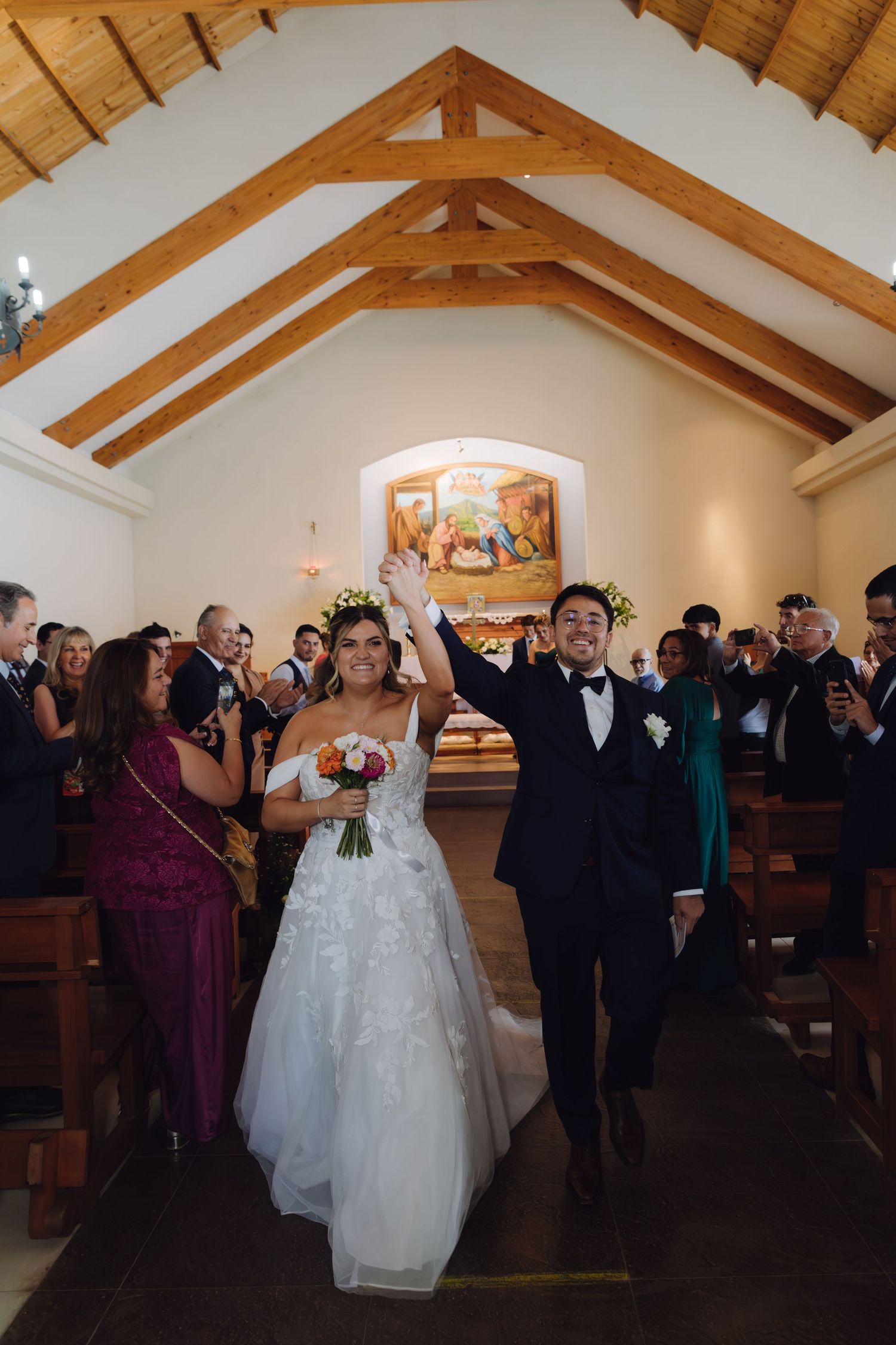 Bride and groom raise hands triumphantly while exiting church aisle surrounded by cheering wedding guests.